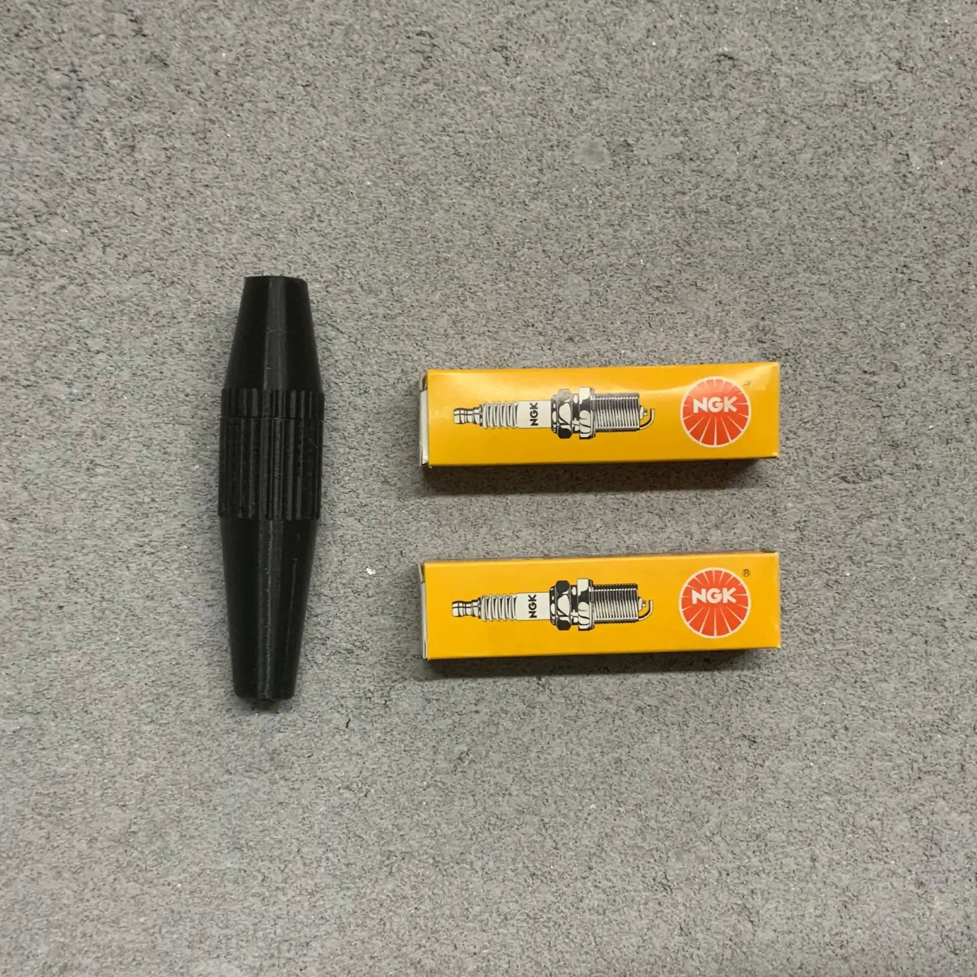 Two NGK spark plugs in yellow boxes with a black spark plug on a gray surface