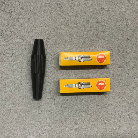 Two NGK spark plugs in yellow boxes with a black spark plug on a gray surface